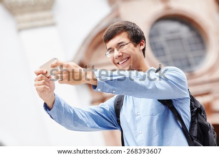 Smiling young man in glasses taking photo of himself with smartphone near cathedral during vacation