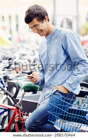 Cheerful young man in glasses and blue shirt with hand in pocket typing message by mobile phone leaning on railing on bicycle parking