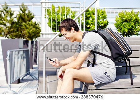 Young joyful man in glasses with backpack and mobile phone sitting on bench and reading message