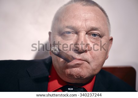 Close up portrait of serious middle aged businessman wearing black suit and red shirt smoking cigar in office