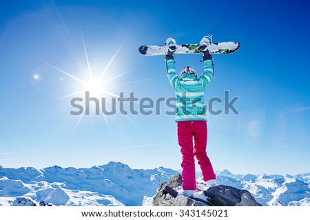 Back view of female snowboarder wearing colorful helmet, blue jacket, grey gloves and pink pants standing with snowboard raised overhead and enjoying sunnymountain landscape - winter sports concept
