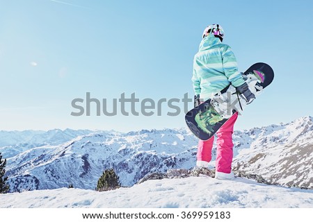 Back view of female snowboarder wearing colorful helmet, blue jacket, grey gloves and pink pants standing with snowboard in one hand and enjoying alpine mountain landscape - snowboarding concept