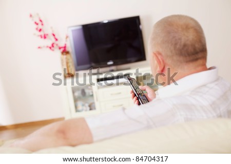 Middle aged man in home bright living room sitting on comfortable couch and watching TV
