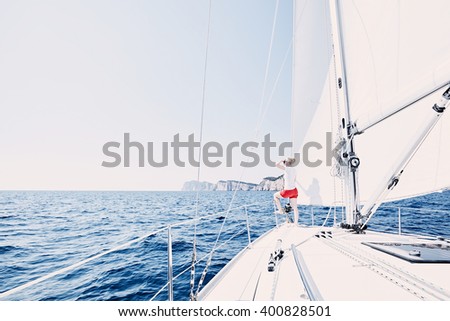 Young woman wearing sunglasses and red shorts, standing on deck under sails on yacht bow, enjoying wonderful view to peaceful sea looking far covering her eyes by hand during summer sailing holidays