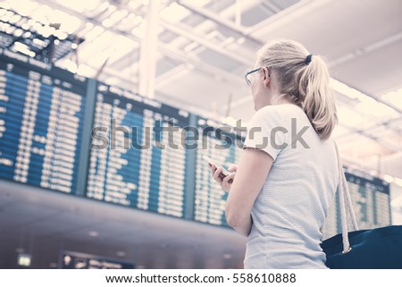 Young blonde woman with phone in her hand and shoulder bag checking flight timetable in international airport - travel concept