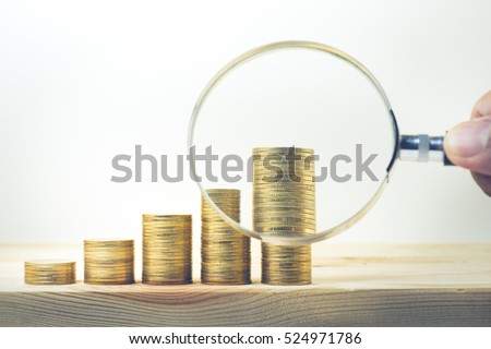 Male hand holding magnifying glass with coin stacked on wood table.For business growth and financial concept