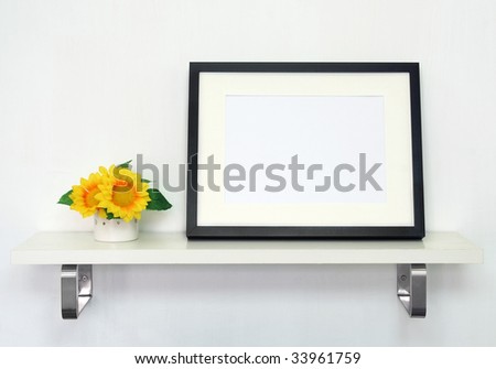 Home Decoration Picture Frame. Potted sunflowers and a black picture frame on white shelf against a white wall.