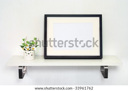 Home Decoration Photo Frame. Potted daisies and a black photo frame on white shelf against a white wall.