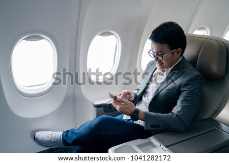 Young asian businessman with suit sitting in business first class seat inside airplane via using smartphone with happiness