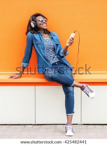 Beautiful smiling african woman with headphones listens to music over orange background.  Fashion woman in sunglasses outdoor.