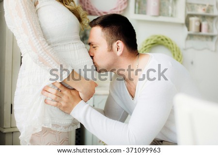 Close up of a happy man kissing the belly of his lovely pregnant wife standing in the white bedroom.  Couple dressed in white clothes. Dad waits the birth of the baby. Sensual photo.