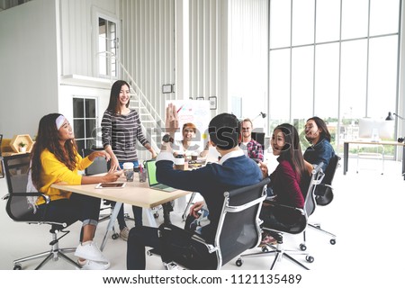 Group of multiracial young creative team talking, laughing and brainstorming in meeting at modern office concept. Female standing and man raising hand for sharing while sitting together in rear view.