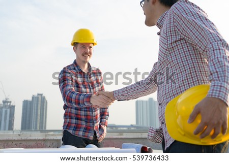 Architect engineer shaking hands other hand he is holding a helmet at construction site against clear sky. With blueprint on table. - business teamwork, cooperation, success collaboration concept