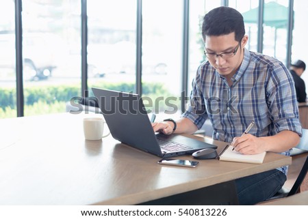 Young male businessman writing on a notepad in front of a laptop on wood desk in coffee cafe shop. Student using notebook at college.
