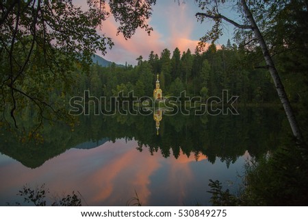 A small Christian chapel stands on the shore of forest lake. Her beautiful silhouette reflected in the mirror water.