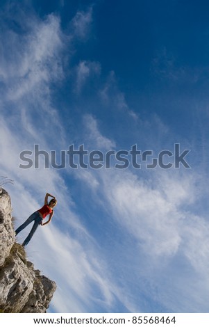 A young girl stands on the edge of a cliff against the sky with clouds