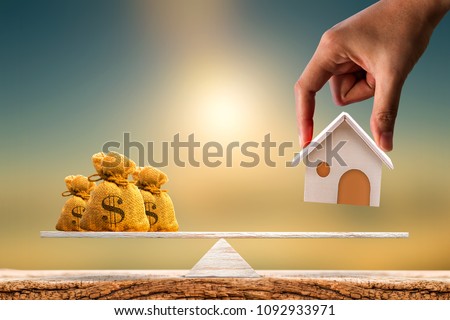 Woman hand hold a wooden home model and money bag put on the scales with balance put on the wood on sunlight, Saving for buy a new house or real estate and loan for plan business investment concept.