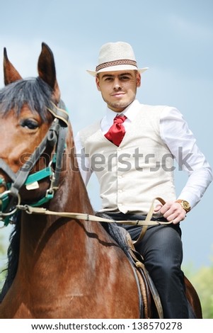 Young stylish man with tie and hat riding a horse on countryside with cloudy background