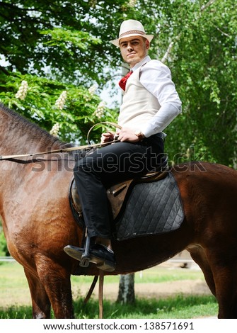 Young stylish man with tie and hat riding a horse on countryside