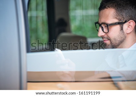Man with glasses sitting in a car backseat looking at a mobile phone