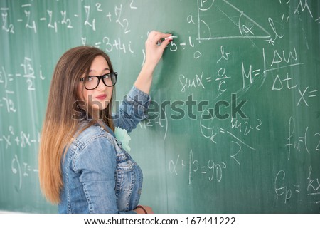Cute schoolgirl with glasses posing solving math problem on blackboard