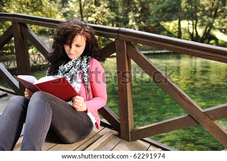 Young beautiful girl student sitting on wooden bridge over the river  in nature and reading a book