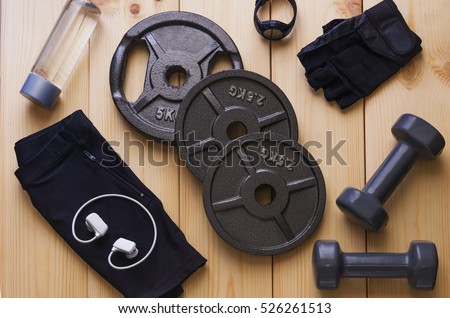 Top view of accessories for fitness in black grey tone. Dumbbells, weight plates,  gloves for gym, sport watch,music player and bottle of water on wooden background. Concept for sport or workout.