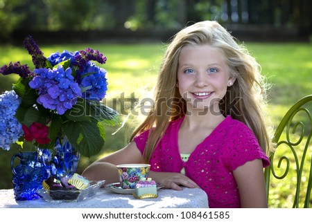 Beautiful little blue-eyed, blonde girl sitting at table with flowers and tea