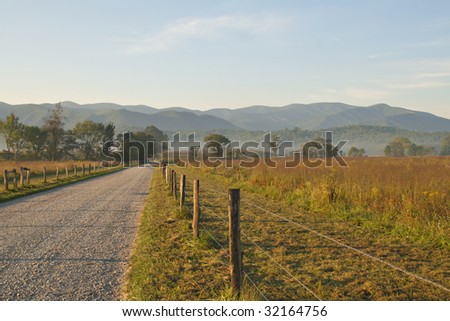 Beautiful un-paved country road lined with trees and fence.  Golden sunrise light on the area and hazy mountains are shown.
