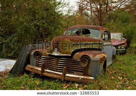 Junk yard vehicles showing old rusted car in overgrown weedy area