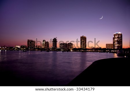 Sunset on the waterfront in St. Petersburg, Florida showing buildings, boats, bay and heron bird in shillouette and cresent moon.  The sky and water shows shades of purple and orange.