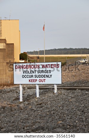 Sign at Guntersville Dam on the Tennessee River indicating Dangerous Waters, Violent Surges Occur Suddenly, Keep Out.  The hyroelectric dam and American flag are in the background.