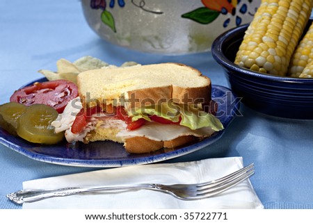 Turkey Sandwich with lettuce and tomato, pickles, chips and corn.  A pitcher of lemonade in the background and fork and napkin in the foreground.  A bite of the sandwich is missing.