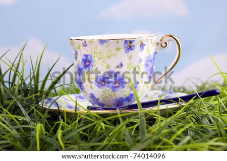 Beautiful colorful ornate coffee or tea cup and saucer sitting in grass with blue sky and clouds in background
