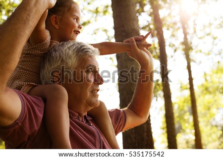Grandfather giving granddaughter piggyback ride in park. Granddaughter sitting on shoulder of older man.