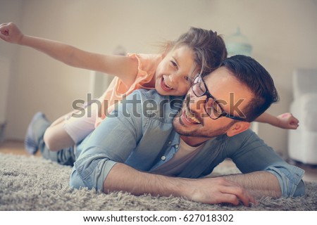Father and daughter spending time at home. Little girl lying on fathers back with open arms. Looking at camera.