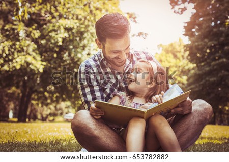 Single father sitting on grass with little daughter and reading book story. Little girl sitting on father lap.