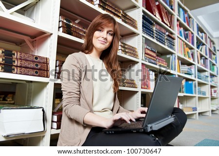 Studying young teenage college student girl  in a library with books and laptop in front of book shelves