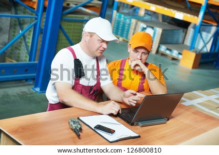 two young handsome workers man in uniform in front of warehouse rack arrangement stillages using notebook laptop computer
