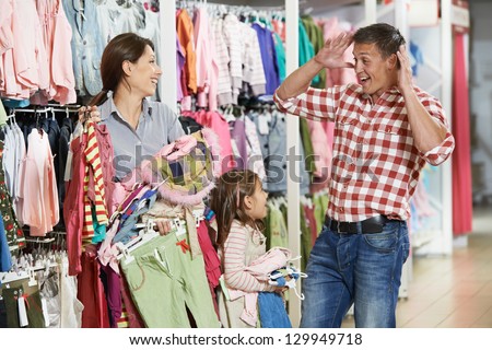 Family shopping. Woman and girl holding clothes with highly surprised father man during shopping at outerwear supermarket