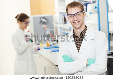 Portrait of young smiling male researcher in front of female researcher carrying out scientific test in chemistry laboratory