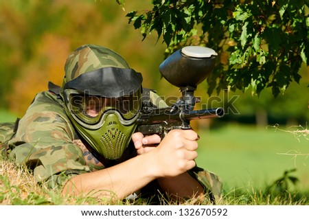 paintball player in protective uniform and mask aiming and shooting with paint marker gun outdoors