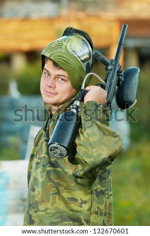Happy paintball sport player man in protective camouflage uniform and mask with marker gun outdoors