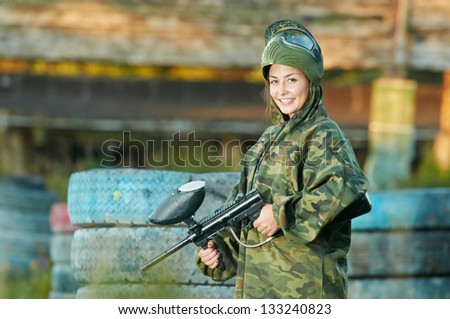 Happy paintball sport player girl in protective camouflage uniform and mask with marker gun outdoors