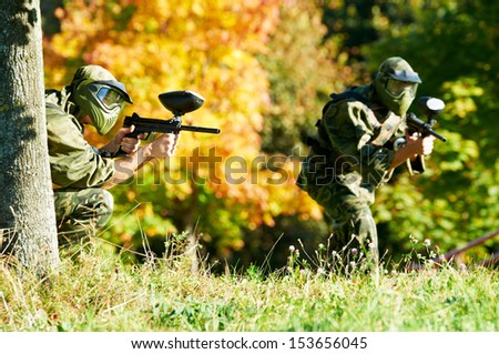 Two Paintball Sport Players In Prootective Uniform And Mask Aiming And Shoting With Gun Outdoors