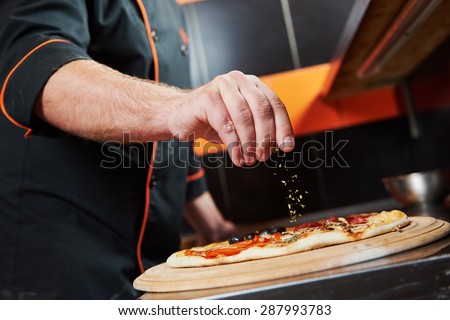 hand of chef baker in uniform adding spice into pizza after pizza preparation at restaurant kitchen