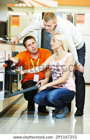 Seller assistant demonstrating cooker stove to young family in home appliance shopping mall supermarket