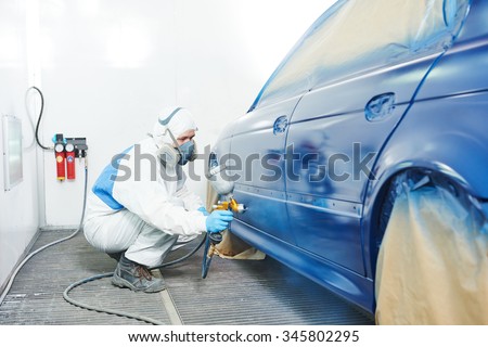 automobile repairman painter in protective workwear and respirator painting car body bumper in paint chamber