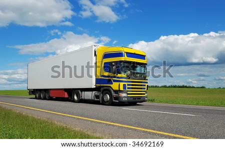 Truck (blue-yellow cabin) and white trailer moving on highway over bright sky.