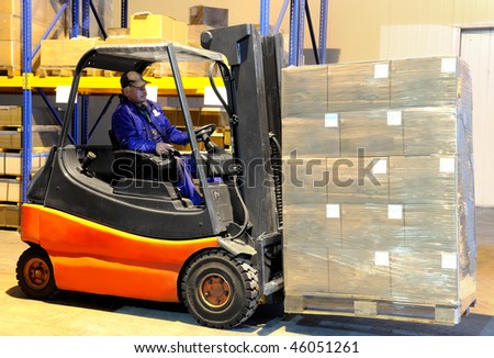 Worker driver of a forklift loader in blue workwear at warehouse with cardboard boxes on pallet